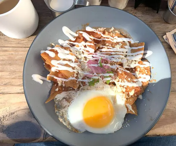 Bu resim canlı bir tabak chilaquiles sergiliyor, geleneksel bir Meksika kahvaltı tabağı. Çıtır tortilla cipsi, üzerinde kremalı beyaz bir çiseleme olan kırmızı bir sosla yıkanır. 