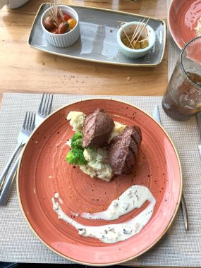 Bu resim, desenli, terakotta renkli bir tabakta güzel kaplanmış bir gurme tabağı sergiliyor. Ana yemek taze et dilimleri gibi görünüyor, muhtemelen sığır eti.