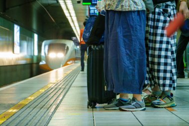 Düşük açılı perspektif, muhtemelen Tayvan 'da, modern bir tren istasyonunda sabırla bekleyen bavullu yolcuları yakalıyor. Arka planda, hafif mot gösteren, farları açık bir şekilde yüksek hızlı bir tren yaklaşıyor.