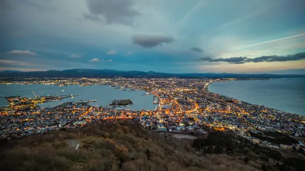 Hakodate Dağı gözlem güvertesinden gün batımında panoramik görüntü Hakodate 'nin şehir ışıklarını parlamaya başlarken yakalıyor. Gökyüzü turuncudan maviye dönüşüyor, büyülü bir sahne yaratıyor..