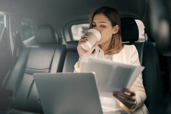 Young woman sitting in the back seat of the car with a paperwork in her hand and drinking coffee.