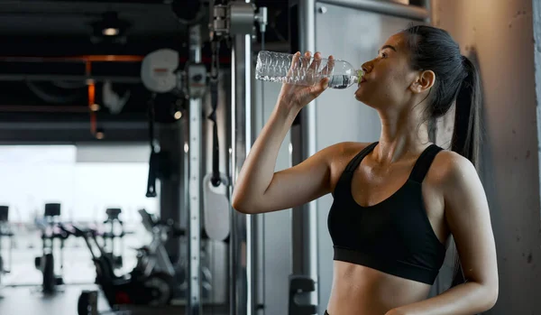 stock image Young woman drinking water from bottle, taking break from exercise at gym.