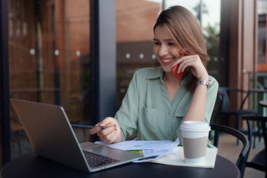 Genç iş kadını kahve dükkanında masada oturuyor cep telefonuyla konuşuyor ve masadaki dizüstü bilgisayara bakıyor..