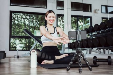 Asian woman training exercise on yoga mat and using mobile phone. Female sport blogger recording video by smart phone. Exercise online training class at gym indoors.