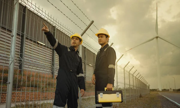 Two technician engineer in uniform with standing and checking wind turbine power farm power generator Station. Clean energy and environment.