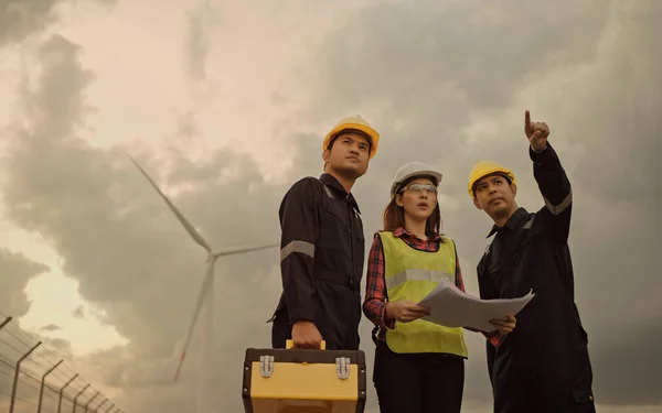 Three technician engineer in uniform with standing and checking wind turbine power farm power generator Station. Clean energy and environment.