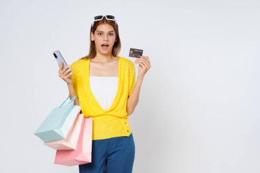 Kredi kartı ve cep telefonu kullanan, beyaz arka planda izole edilmiş alışveriş torbaları taşıyan heyecanlı genç bir kadın..