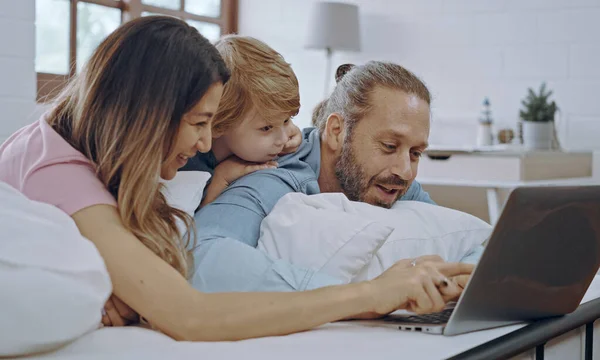 stock image Caucasian family of three using laptop while lying on bed together, browsing internet or watching movie.