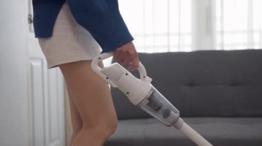 Genç bir kadın modern beyaz oturma odasında kablosuz elektrik süpürgesiyle yerleri temizliyor..