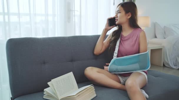 自宅でソファーで電話を使用して腕を痛めるために柔らかいスプリントを着用している若いアジアの女性 — ストック動画