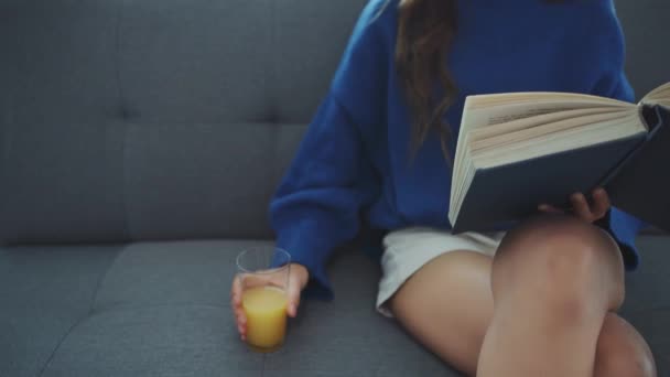 年轻的女人喝了一杯橙汁在家里放松一下 她坐在沙发上 在家看书 — 图库视频影像