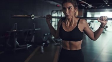 Genç bir kadın spor salonunda sırtında halterle çömeliyor..