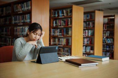 Stresli genç kız öğrenci tahta masada oturuyor ve kütüphanede sınav hazırlıkları sırasında kitap okuyor..