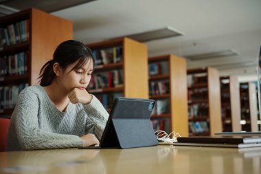 Stresli genç kız öğrenci tahta masada oturuyor ve kütüphanede sınav hazırlıkları sırasında kitap okuyor..