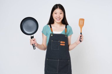 Mutfak önlüğü giyen genç bir kadın yemek pişiriyor, tava tutuyor ve beyaz arka planda izole edilmiş spatula tutuyor..
