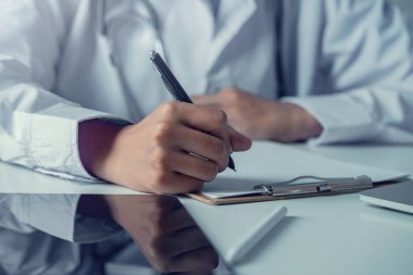 Tıbbi kayıtları dolduran pratisyen hekimin elleri. Beyaz önlüklü doktor iş yerinde dizüstü bilgisayarla evrak işleriyle uğraşıyor, notlar yazıyor, evrak hazırlıyor, raporlar hazırlıyor, reçete yazıyor. Kapat.