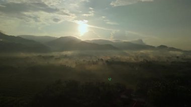 Dağlık bir arazide, yoğun sis örtüsüyle gün doğumunun havadan görünüşü. Güneşin altın ışınları sisi delip geçer ve tüm sahnenin üzerine huzur dolu bir parıltı saçar..