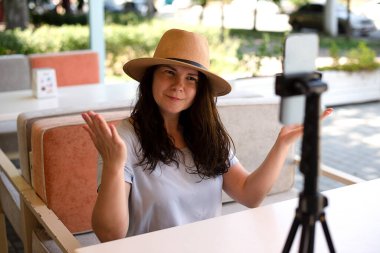 Attractive smiling woman outside sitting with tripod and smartphone,making video call or providing online lesson.