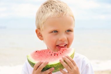 Tatlı güneş yanığı sarışın çocuk sahilde, elinde sulu bir karpuz dilimi tutuyor ve onu yiyor..