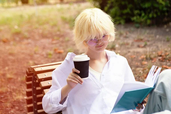 Teenage girl with blond hear drinking coffee from craft paper cup outside and reading book.