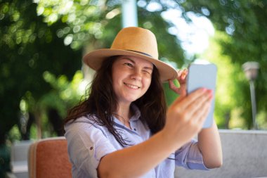 Stylish woman in hat with smartphone outside winking and smile.