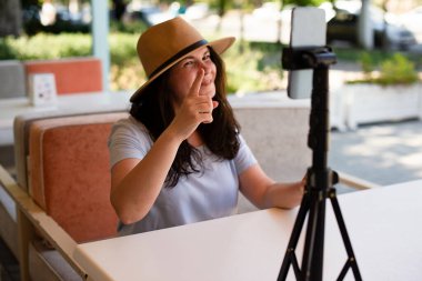 Attractive smiling woman outside sitting with tripod and smartphone,making video call or providing online lesson.