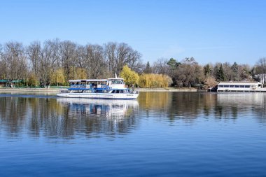 Bucharest, Romania, 21 November 2021: Landscape with white boat on Herastrau lake and large green trees in King Michael I Park (Herastrau) in Bucharest, Romania, in a sunny autumn day