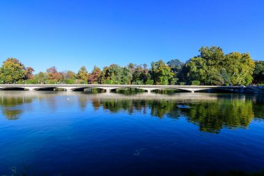 Romanya 'nın başkenti Bükreş' teki Carol Park 'ta güneşli bir sonbahar gününde, uzun gri köprüsü ve göl kenarındaki çok sayıda büyük yeşil ağaçla manzara