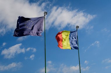 Avrupa Birliği ve Rumen ulusal bayrakları güneşli bir bahar gününde bulutlu mavi gökyüzüne doğru rüzgarda dalgalanıyor