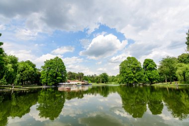 Craiova, Romanya, 28 Mayıs 2022: Dolj ilçesindeki Nicolae Romaescu parkında göl, suluzambaklar ve güzel güneşli bir bahar gününde mavi gökyüzü ve beyaz bulutlarla dolu büyük yeşil treyler ile canlı manzara