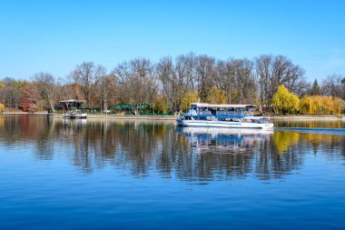 Bucharest, Romania, 21 November 2021: Landscape with white boat on Herastrau lake and large green trees in King Michael I Park (Herastrau) in Bucharest, Romania, in a sunny autumn day