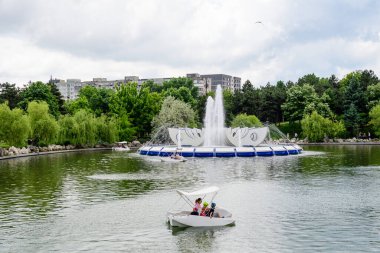 Bükreş, Romanya - 25 Mayıs 2021: Bulutlu bir yaz gününde, Drumul Taberei Parkı 'nda (Parcul Drumul Taberei) modern beyaz su çeşmesi, göl ve yeşil ağaçlarla çevrili manzara
