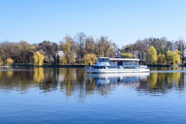 Bucharest, Romania, 21 November 2021: Landscape with white boat on Herastrau lake and large green trees in King Michael I Park (Herastrau) in Bucharest, Romania, in a sunny autumn day