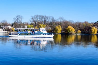 Bucharest, Romania, 21 November 2021: Landscape with white boat on Herastrau lake and large green trees in King Michael I Park (Herastrau) in Bucharest, Romania, in a sunny autumn day