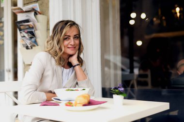 Küçük bir kafe masasında oturan bir kadın lezzetli bir salata ve ekmeğin tadını çıkarırken gülümsüyor. Atmosfer doğal ışık ve şehir hayatı ile dolu..