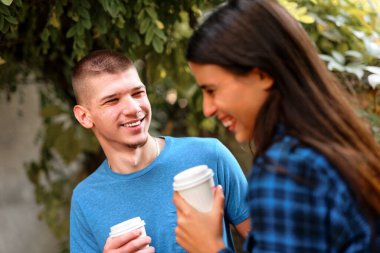 Neşeli genç bir adam ve kadın parkta ellerinde kahve fincanları ile duruyorlar. Gülüşleri ve kahkahaları paylaşırlar. Etrafları yeşilliklerle çevriliyken güneşli bir günün tadını çıkarırlar..