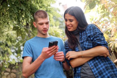 İki genç arkadaş, güneşli bir günde çevrenin yeşillikle çevrili olduğu bir günde akıllı bir telefona bakarken hareketli bir sohbete dalarlar..