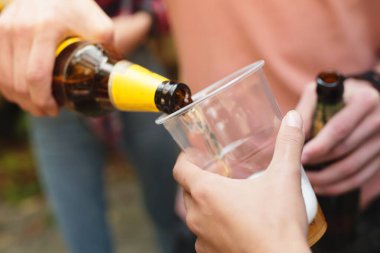 Canlı bir açık hava toplantısında, bir kişi açık bir bardağa içki dökerken, diğeri seyrederek arkadaşlar arasında rahat ve şenlikli bir atmosfer yaratıyor..