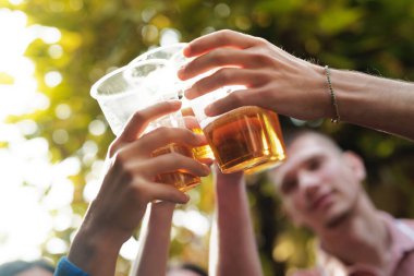 Bir grup arkadaş, canlı bir bahçede, sıcak güneşin altında birbirlerine eşlik edip gülümseyerek kadeh kaldırır ve eğlenirler. Kahkaha havayı dolduruyor..