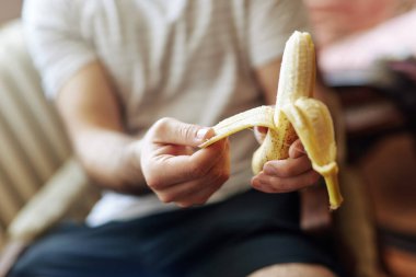 Bir insan rahat bir sandalyede otururken iki eliyle muz soyuyor. Sıcak, davetkar atmosfer sağlıklı atıştırmalıkların rahatlamasını ve eğlenmesini öneriyor..