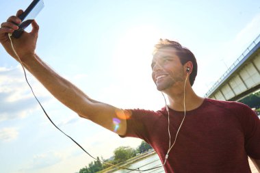 Genç bir adam nehir kenarında durur, selfie çekerken geniş bir gülümsemeyle. Kulaklık takıyor ve elinde akıllı bir telefon tutuyor. Arka planda güneşli güzel bir gün sergiliyor..