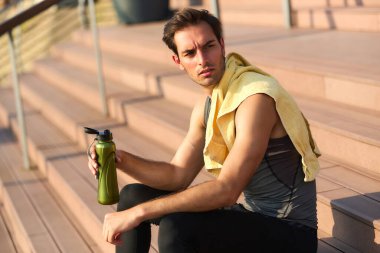 Genç bir adam ahşap merdivenlerde oturur, yoğun bir egzersizden sonra soluklanır. Omzuna sarılı bir havlusu var ve sıcak güneş ışığının tadını çıkaran bir su şişesi tutuyor..