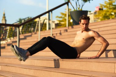 Üstsüz bir adam şehir parkında çelik ve ahşap basamaklara triseps dalışı yapar. Ilık güneş ışığı, hareketli bir akşam atmosferi yaratıyor, çalışmalarını vurguluyor..