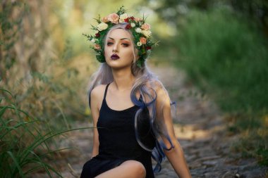 Çiçekli bir taç ve çarpıcı bir makyajla süslenmiş gotik bir kadın canlı yeşil bir alanda güvenle oturuyor. Yaprakların arasında şık bir şekilde poz verirken güneş ışığı onun özelliklerini vurguluyor..
