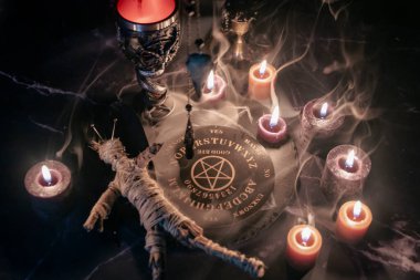 A haunting occult setup featuring a pendulum, mystical symbols, candles, a voodoo doll, and a ritual goblet amidst swirling smoke clipart