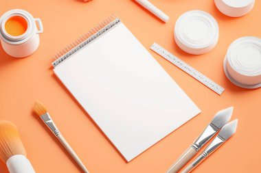 Creative workspace featuring a blank notepad, paintbrushes, and art supplies on a vibrant orange background. clipart