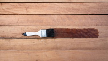 Boya fırçası yeni kereste yüzeyine vernik uygular, üst görünümde ahşap ürünü ve yatay bileşim. Islak, zarif ve koyu kahverengi renkli görünün. Tahıl tabakasını geliştirmek veya kaplamak için..