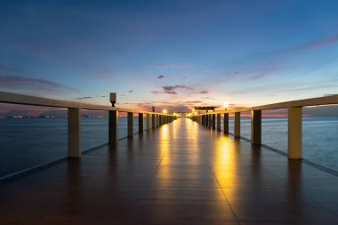 Long walkway floor on wooden structure extending into calm sea at twilight with warm glow of illuminated lamps reflecting on floor. Scenic serene landscape representing tropical seascape background.