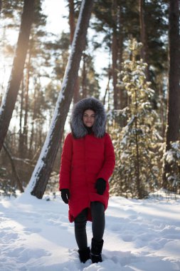 Genç bir kadınla kış ormanında çekilmiş bir fotoğraf. Karlı bir parktaki kız. Donmuş bir nehrin kıyısında kırmızı bir ceketle. Doğada kış yürüyüşü. Soğuk mevsim. Güzel kız, güneşli bir gün..