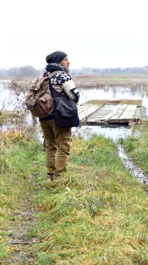 Sırt çantalı genç bir adam. Gezgin, hippi bir göletin kıyısında duruyordu. Su basmış çayırlar. Uzağa bakıyorum, Yürüyüş, Orman, Yolculuk. Dostum, arkadan bak. Doğanın güzelliği
